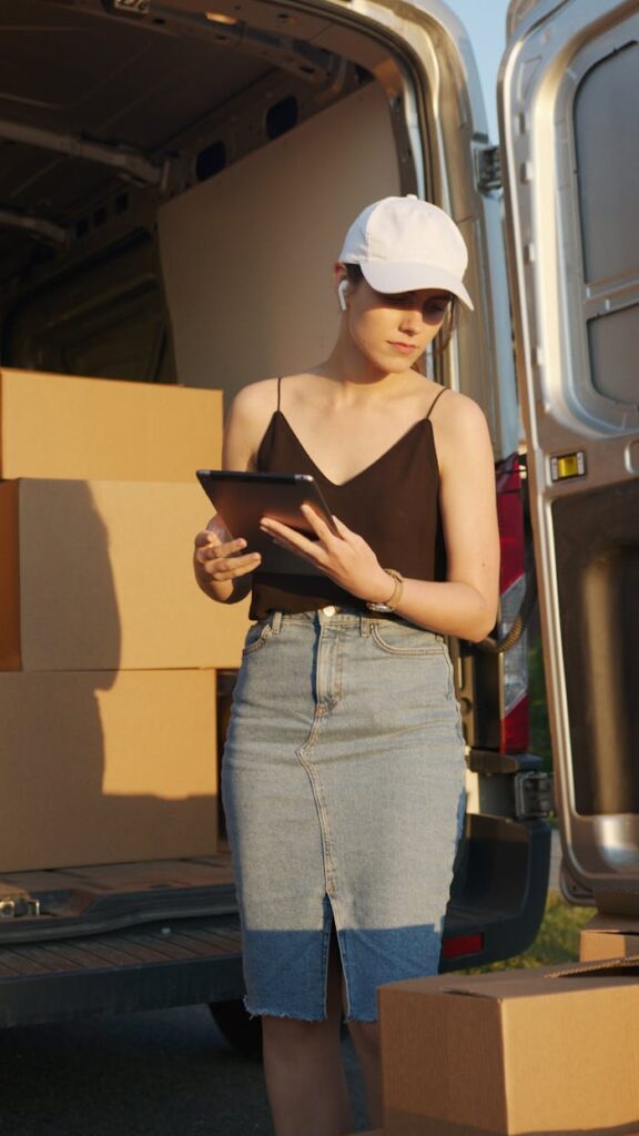 Woman with tablet organizing delivery boxes by open van in warm evening light.
