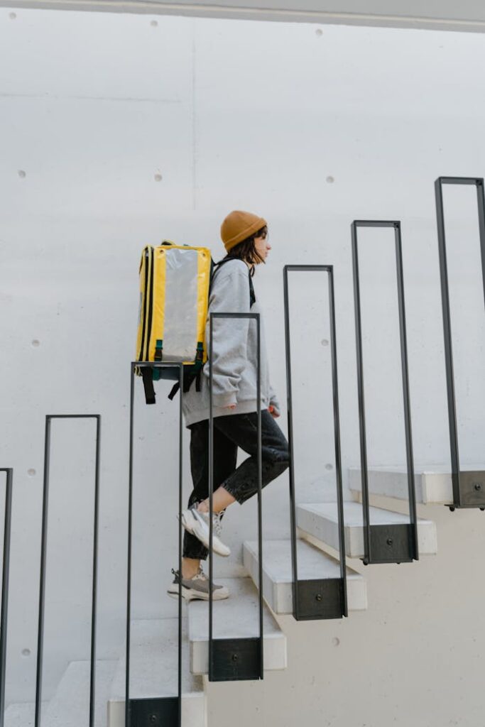 Female courier with yellow backpack delivering food up stairs in urban setting.